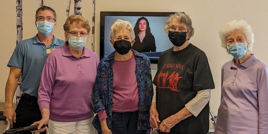 Image of five adults wearing masks and standing in front a TV screen, which is displaying the instructor of an online course. On the left is a middle aged man wearing a uniform and name tag. The other 4 women are dressed casually, and 2 are holding mobility aids.