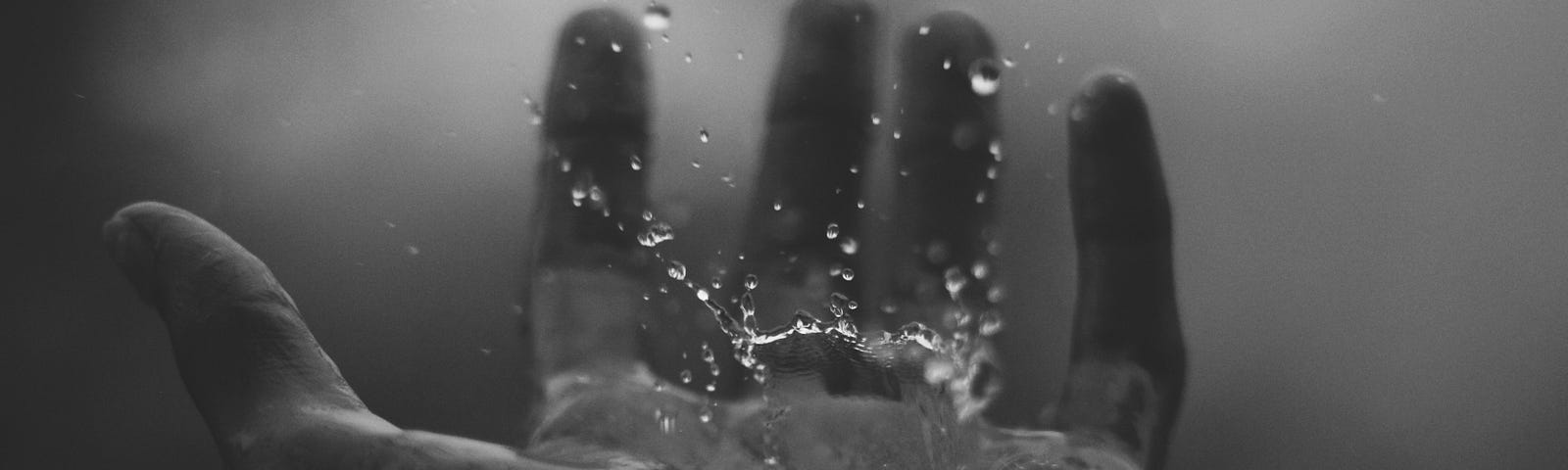 Black and white photo of a left hand, palm up, catch a large raindrop splashing on the palm.
