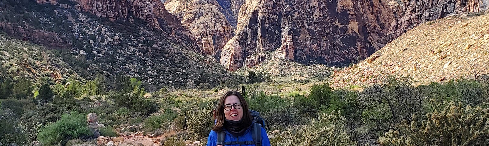 Author in the center of the photo, wearing a blue wind jacket, rust-colored jeans, and hiking boots. In the background, mountains of rocks and a blue sky above.