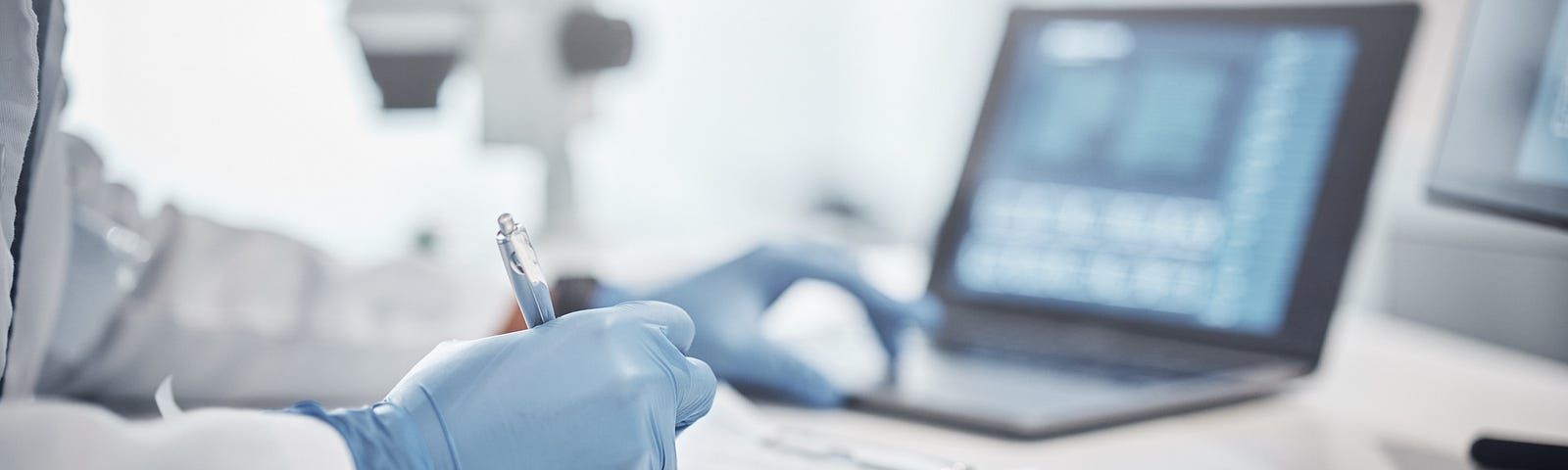Image of a person in a white lab coat and blue gloves taking notes at a laptop