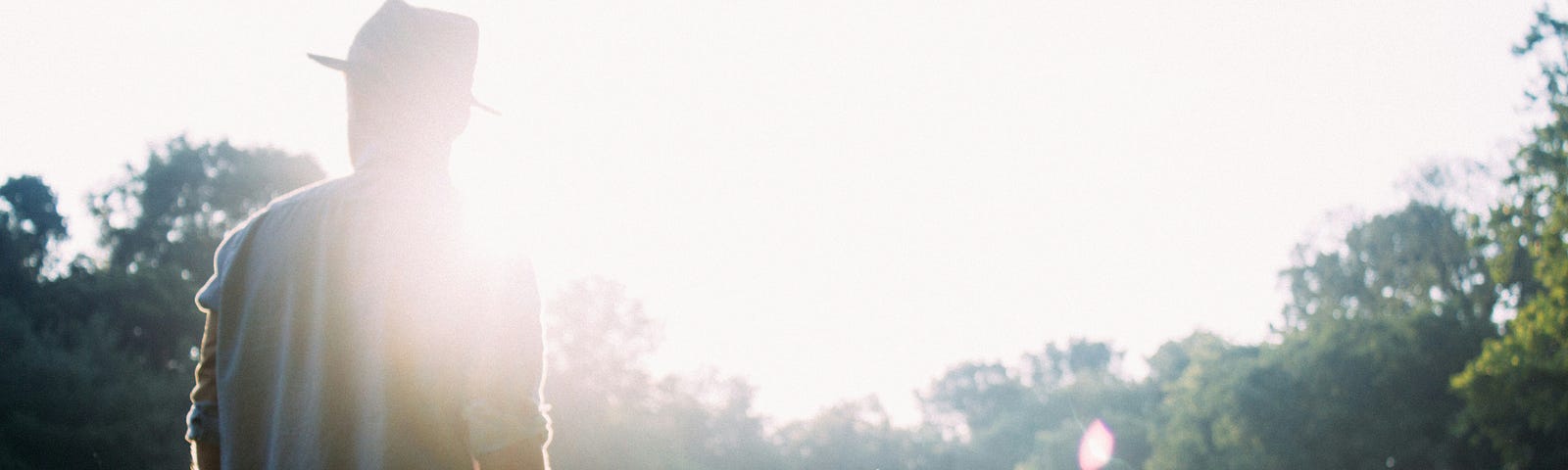 A man standing in a sunlit field, surrounded by trees with rays of sunlight filtering through.