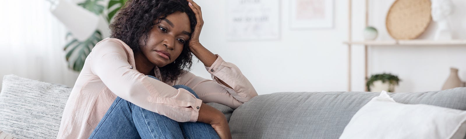 A woman sits on a couch with her knees pulled up to her chest, and her head resting in her free hand.