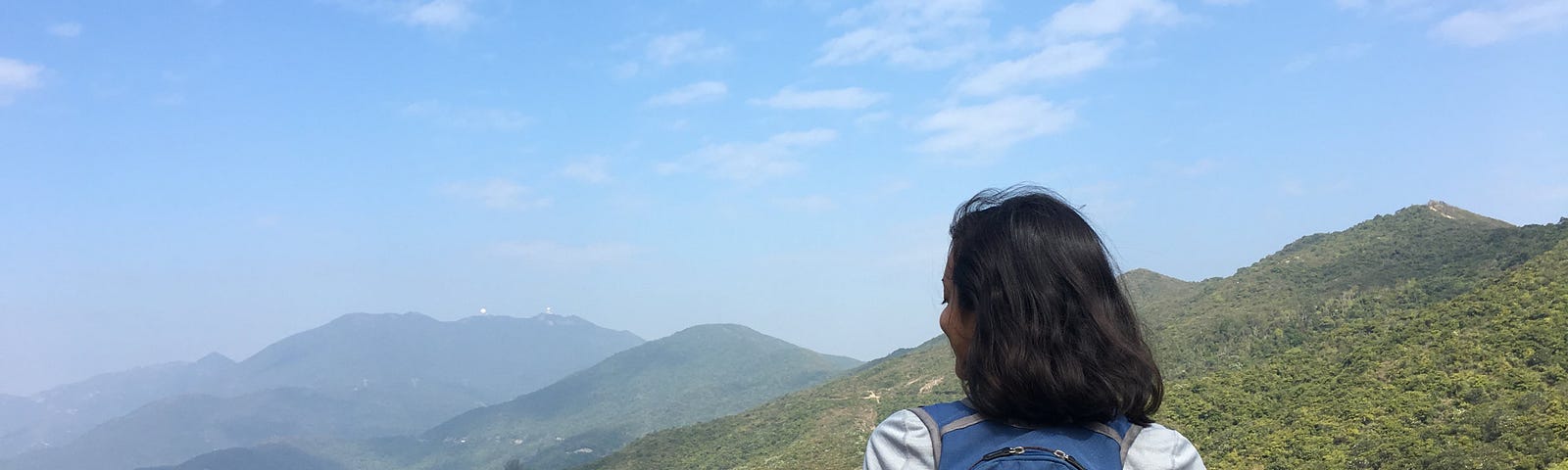 A woman with short dark brown hair is shown from behind. She wears a gray long-sleeved top and blue backpack. She stands in front of a verdant hill, with mountains visible in the distance through haze.
