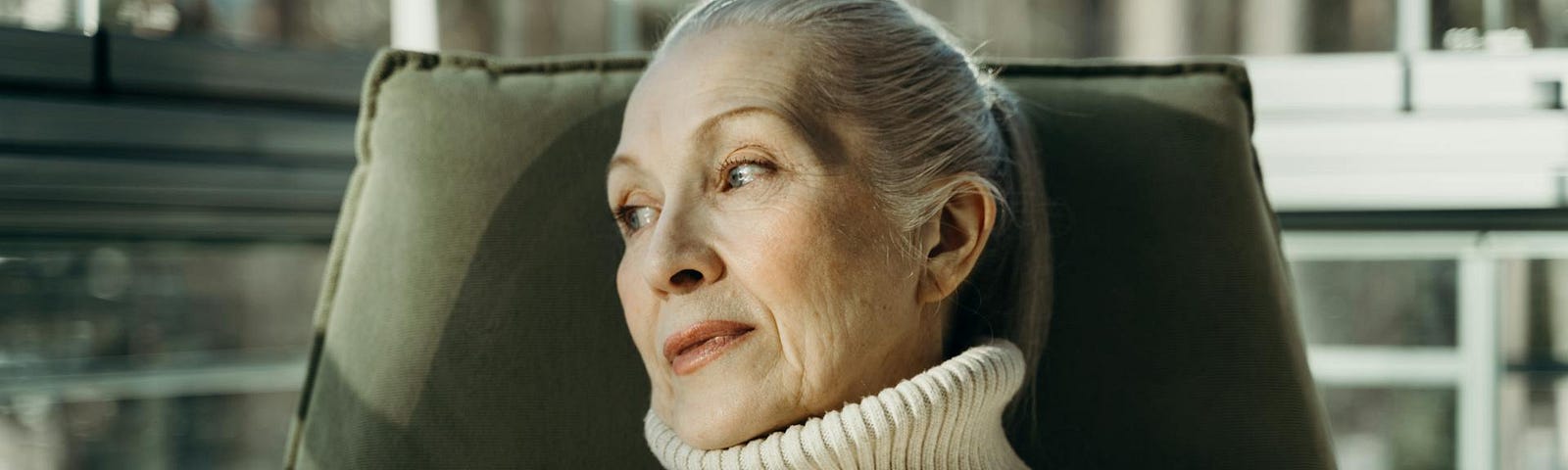An older woman with grey hair pulled back in a ponytail wears a beige turtleneck sweater and glances sideways while sitting in an olive green chair.