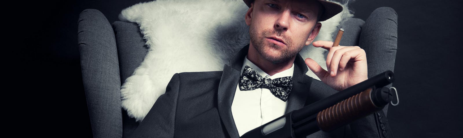 man with mobster appearance wearing old-time hat, bowtie and suit leaning in an armchair, holding a smoky cigarette in one hand and a type of shotgun in the other