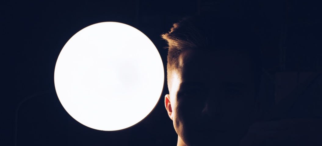 A man’s face is shadowed and standing before a large bright circle of white light.