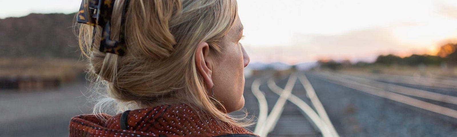 A woman facing away from the camera while standing on train tracks.