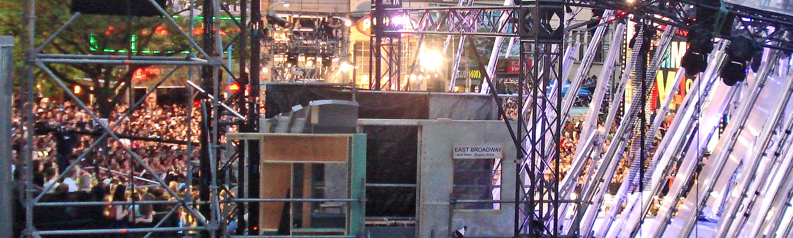 Backstage of an outdoor concert. Scaffolding and a lighting grid are visible in the foreground, stairs lead up to a small room where performers can wait. A huge crowd of people is visible in the background.