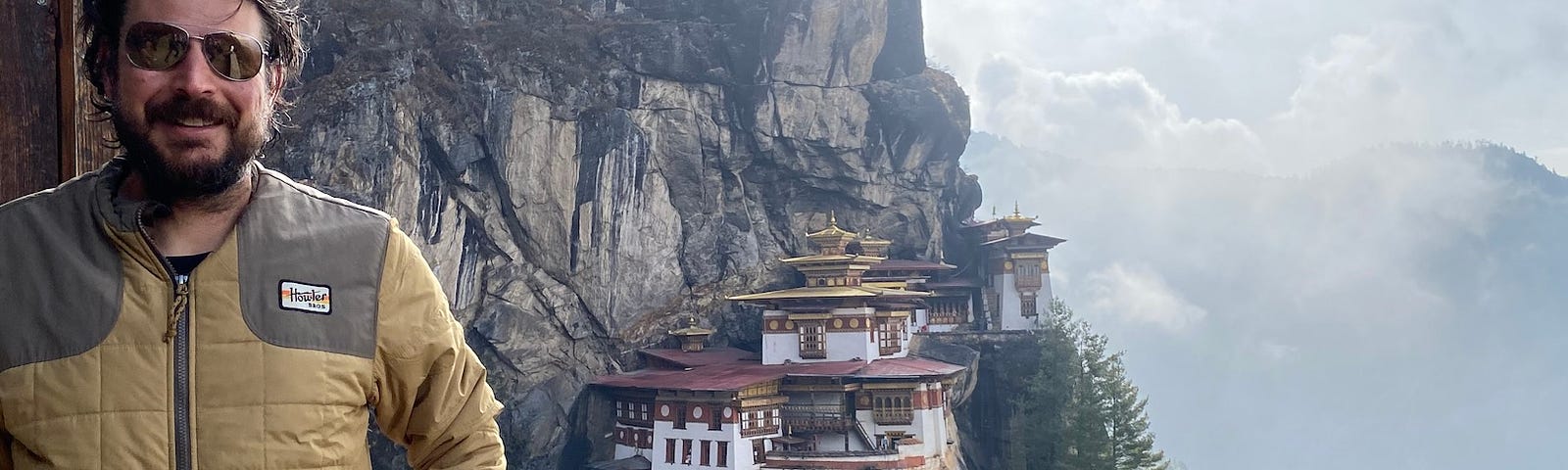 The author, in the foreground, wearing a beige colored warm weather jacket. Tigers Nest Monastery buildings, white with brown roofs, are in the background, clinging to the side of a steep rocky mountain. More clouds and mountains are visible in the distance.