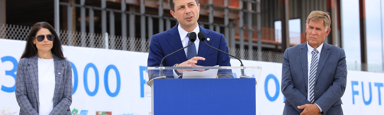 Secretary Buttigieg speaks behind a podium with a sign that says “$8M to Build a Better GRR.” Michigan Attorney General Dan Nessel and Kent County Board of Commissioners Chair Stan Stek stand behind him. The three are in front of a construction project to expand the GRR terminal.