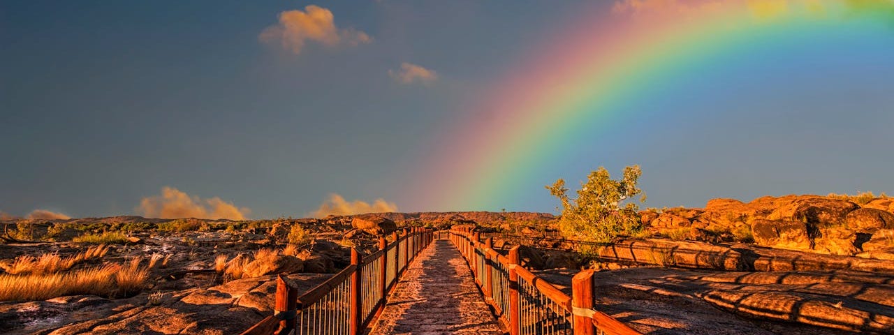 A red wooden walkway with railings heads through a desert-like field headed toward a gorgeous rainbow.