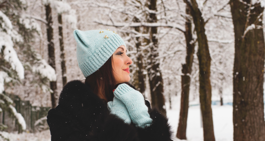 Woman standing in the winter woods in light blue knit hat and gloves. Holing her hands to her heart and smiling.