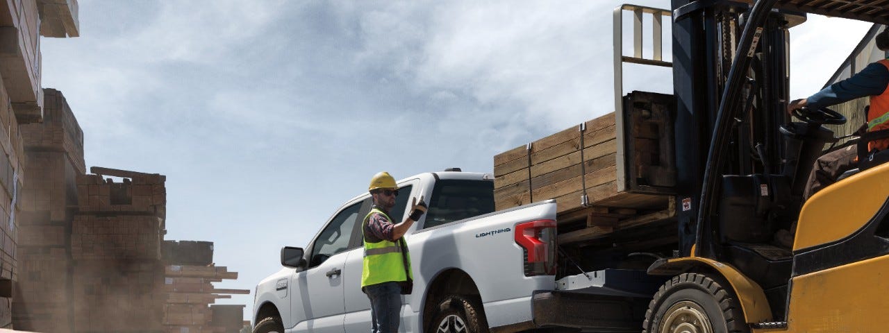 An image of a forklift placing timber onto the back of a new, white fully electric Ford F-150 as a construction worker in a hardhat and safety vest directs the placement.
