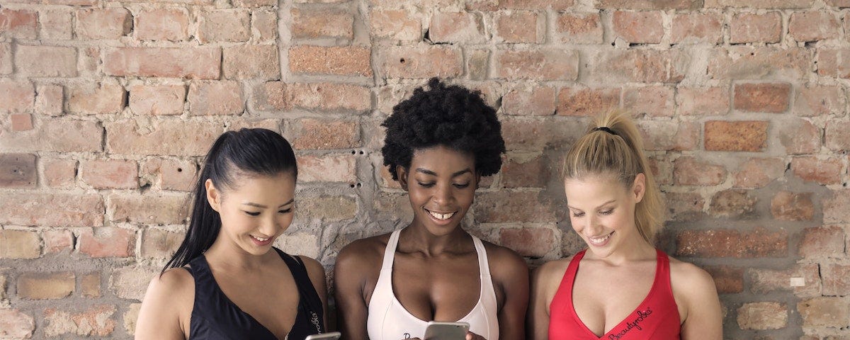 Three women of diverse ethnicity (Asian, African, Caucasian) leaning against an internal brick wall. All three are wearing a sports bra and yoga pants. Each is holding her phone, looking at its screen, and smiling broadly as though amused.