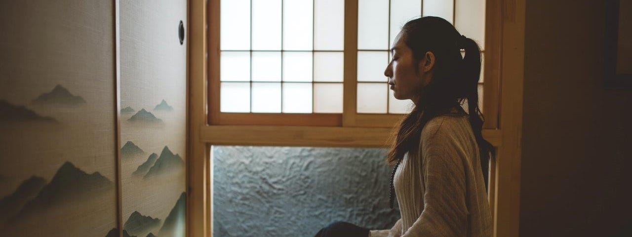 A woman meditating next to a window.