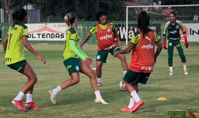 Jogadoras do Palmeiras treinam em Vinhedo. Imagem mostra quatro atletas disputando bola, com camisas verde limão, calção verde escuro e meias brancas, sendo que duas usam colete vermelho. Ao fundo, uma das goleiras com calça e camisa de manga longa verde escuro.