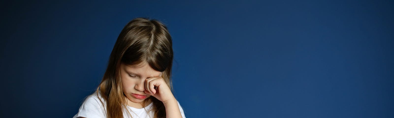 A sad girl hangs her head and rubs her eyes against a deep blue background.