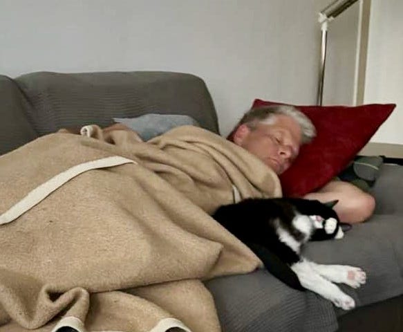 Man asleep on sofa with black and white cat sleeping next to him.