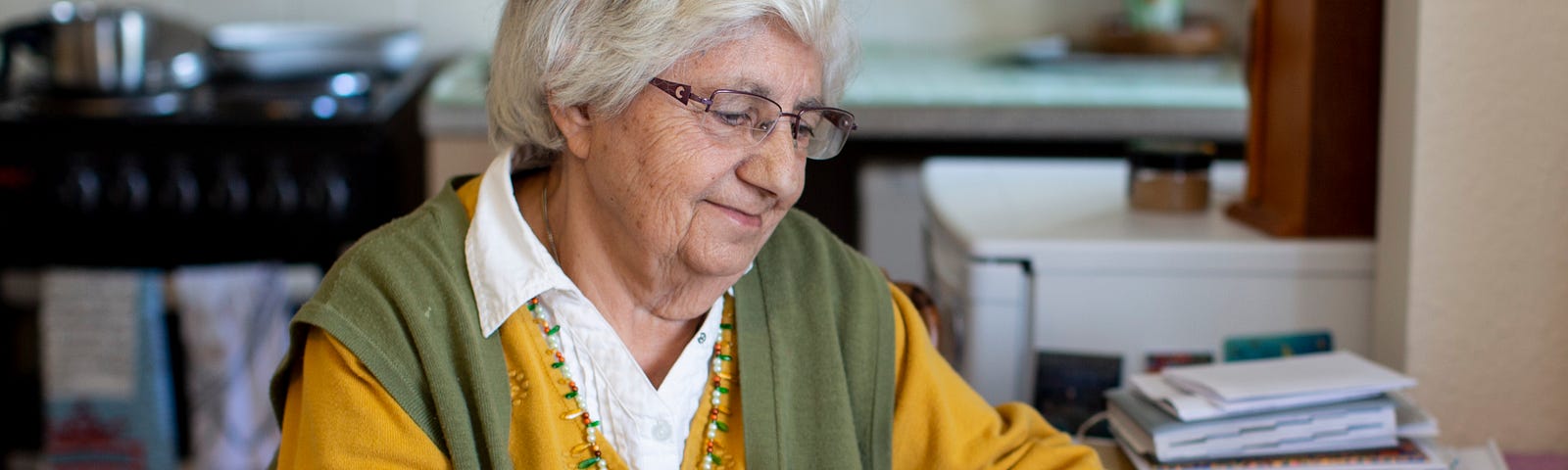 An older woman smiles as she looks through a leaflet while sat at a kitchen table. There are piles of paper next to her.