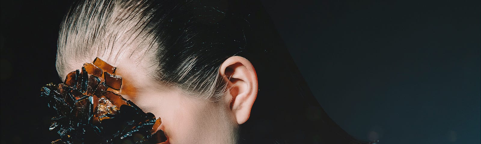 Photograph of a woman. A mass of brown crystals completely obscure her face from the viewer. Her hair is slicked back. The background around her is dark gray.