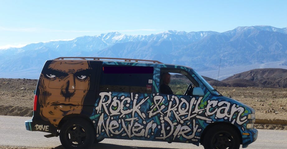 Painted van stopped on the side of a road. there is a man’s face painted near the back of the van with the words “Rock and Roll can Never Die” on the side. Mountains are visible in the distance.
