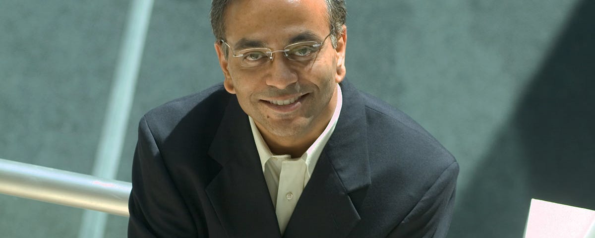 South Asian man in suit and glasses sits with crossed arms, looking up at the camera and smiling