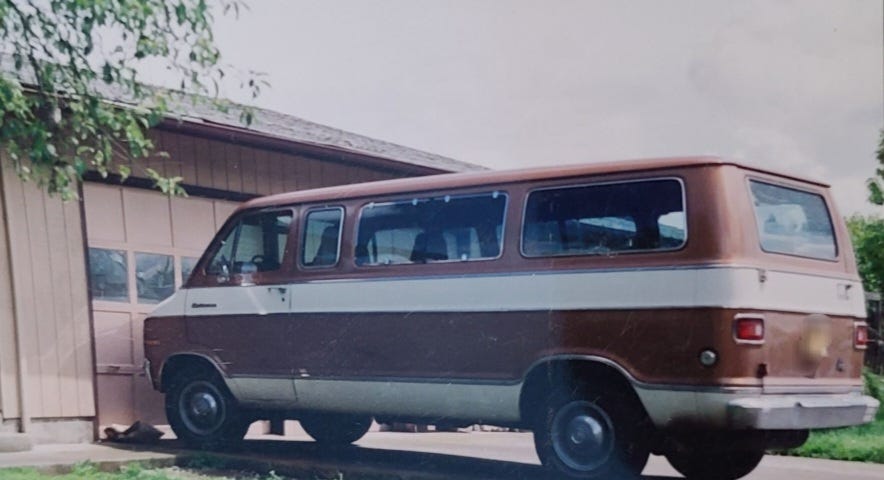 1977 Dodge Van from writer’s childhood, circa 1992, maroon color with white stripes, in front of a tan colored garage door, next to a green lawn, green leaves from tree branches.