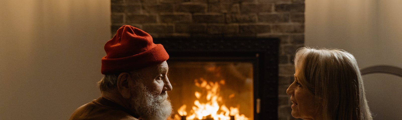 Man in red cap sits in front of a fire with a woman, with long blond hair. They talk.