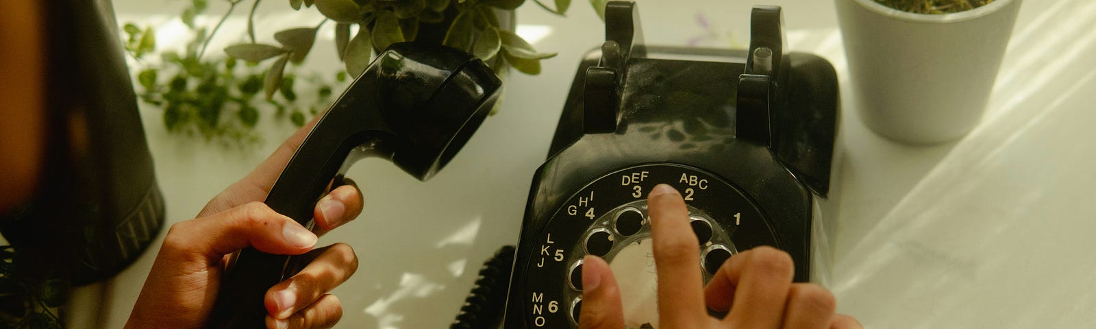 Hands seen using a black rotary phone on a table next to plants.