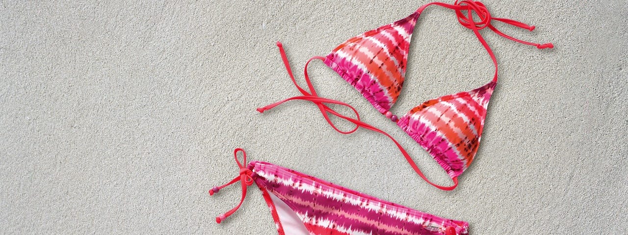 Brightly coloured string bikini, shades of red and pink, and a shell on a sandy background