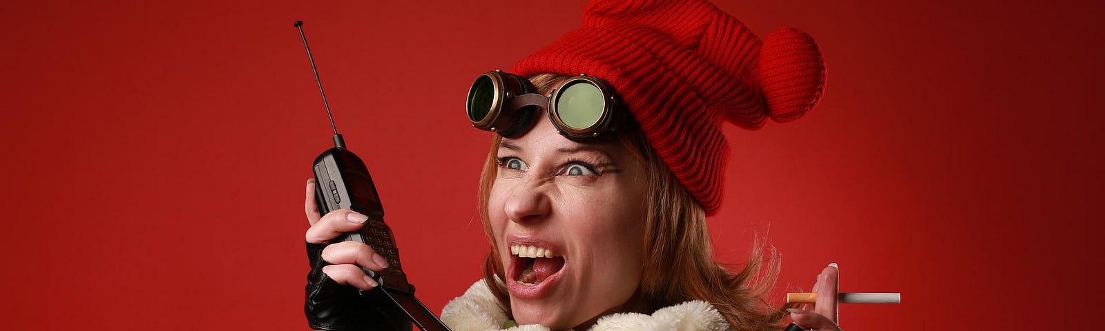 A woman wearing a green winter jacket and a too-small kid’s winter hat is wearing driver’s gloves and vintage motorcycle goggles. She holds a walkie-talkie in her right hand and a cigarette in her left. She has a crazy expression.