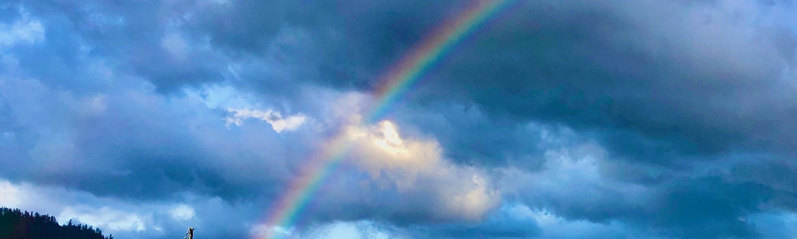 A half rainbow climbs the sky with clouds in the background. There is a hill and the ocean in front with sailboats in the water.