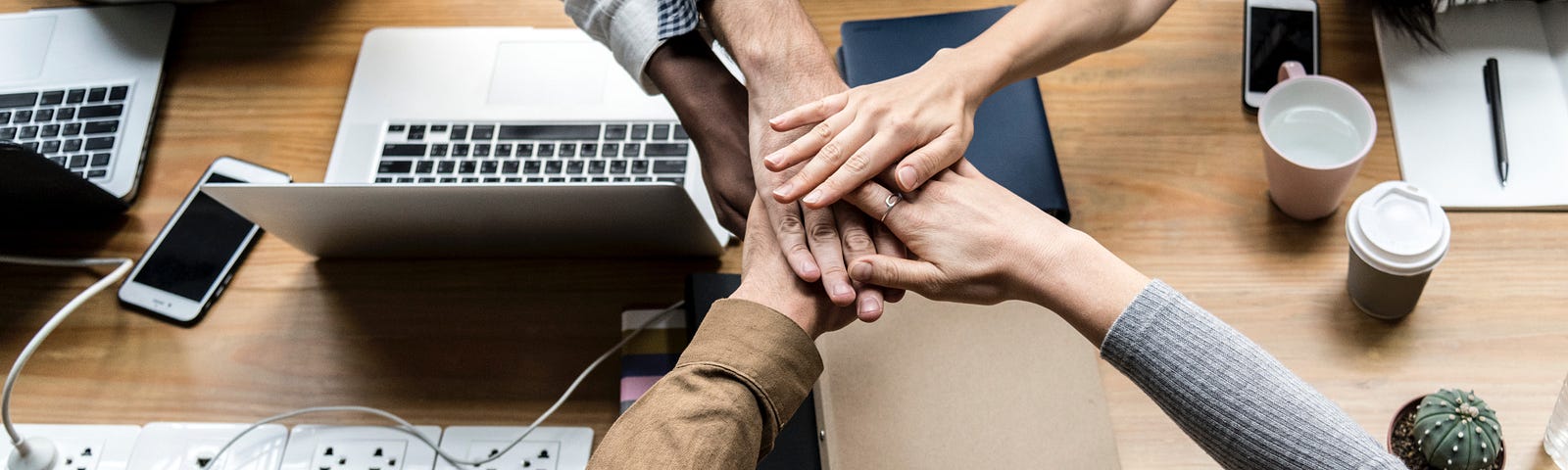 A group puts hands in the middle of the table containing several computers. Crowdsourcing.