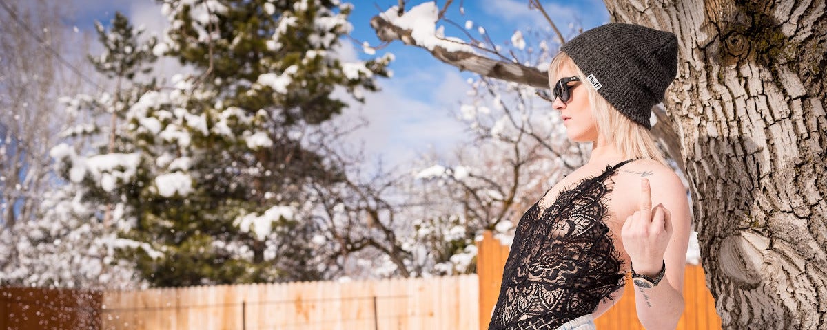 A young, blonde woman wearing unbuttoned denim cut-offs, a black lace body, sunglasses, and a toque. She is leaning against a tree in a back yard, not looking at the camera but raising her middle finger to it. There is snow falling, and laying on the ground. A snow covered tree is visible beyond the yard.