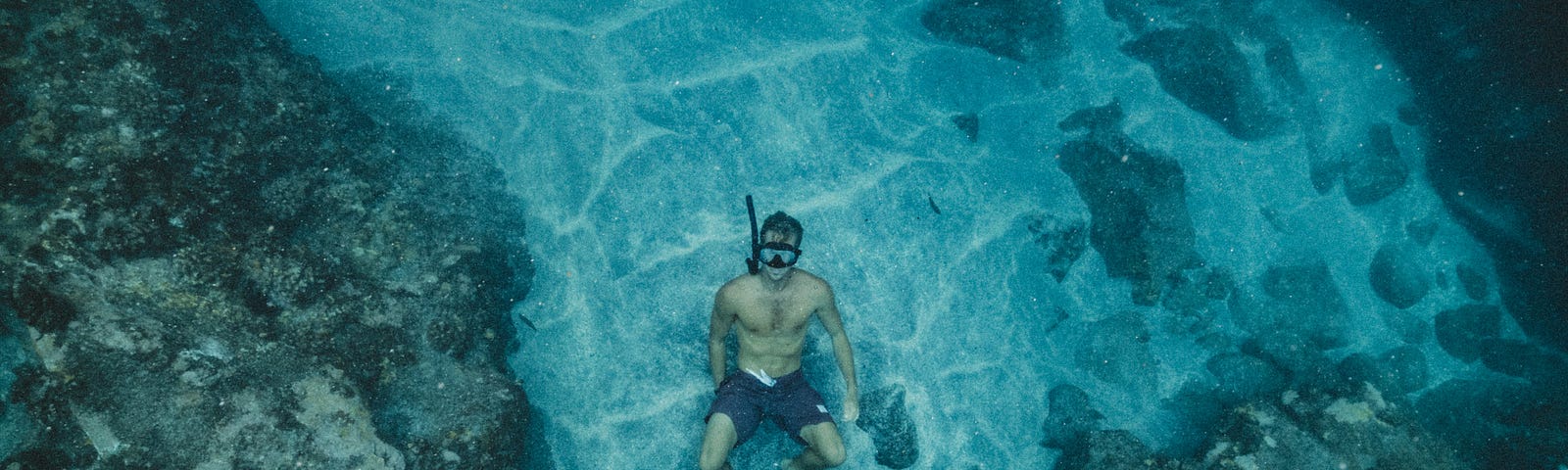 A male snorkeler in swimming trunks at the water’s bottom, leaning back on his knees and feet, on sand which is surrounded by rocky outcroppings.