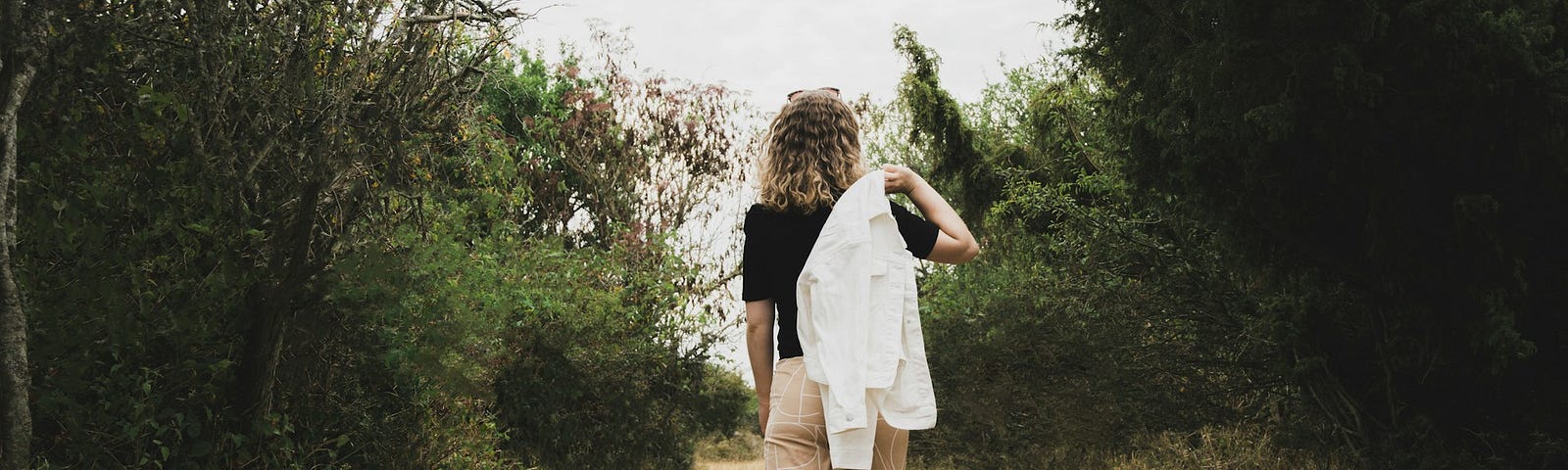 Back of a woman carrying a white dress shirt over her shoulder and walking down a dirt path surrounded by trees.