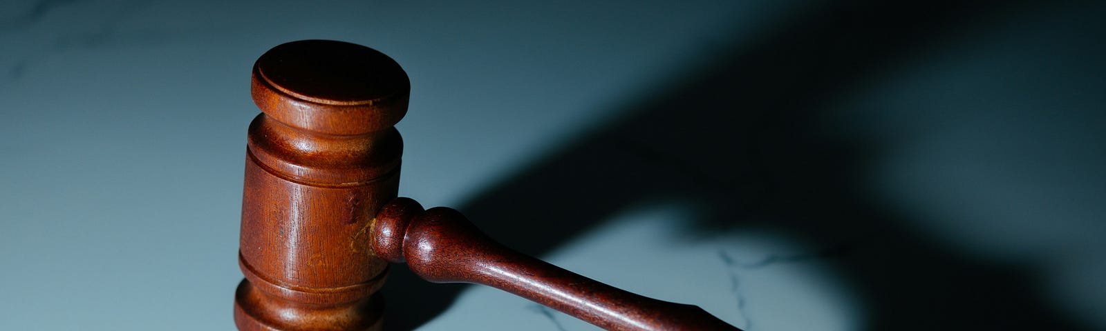 A brown wooden judge’s gavel rests on a light colored marble surface. The gavel’s shadow can be seen at right.