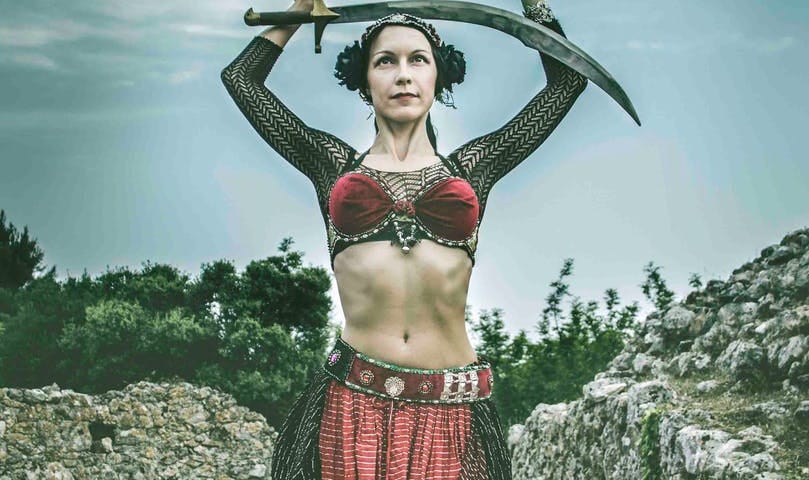 Woman in feminine warrior clothes holds a sword above her head.