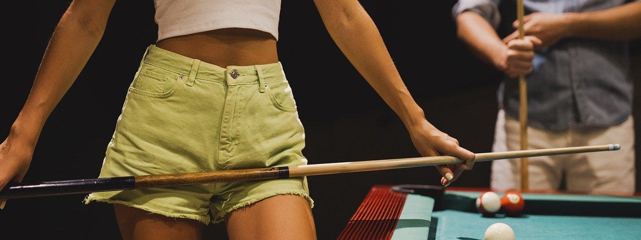 Image shows part of a body wearing a white top and light green shorts, and holding a cue in front of them, with a pool table in the right bottom half of the image.