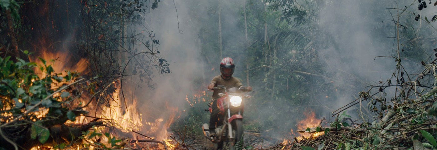 A settler wearing a helmet rides his red motorcycle through a logged, burning patch of Amazon rainforest.