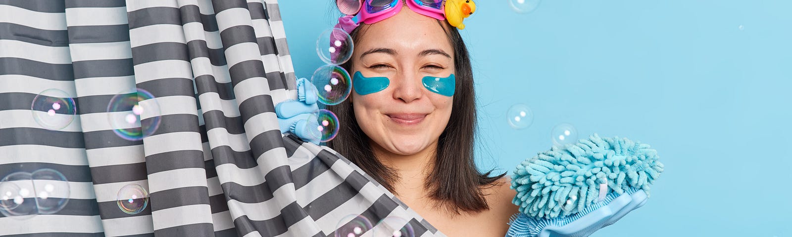 Woman wearing goggles and blue dish gloves with hair in curlers and teal colored face mask under eyes stands in shower with black and white curtain surrounded by bubbles looking content with eyes closed