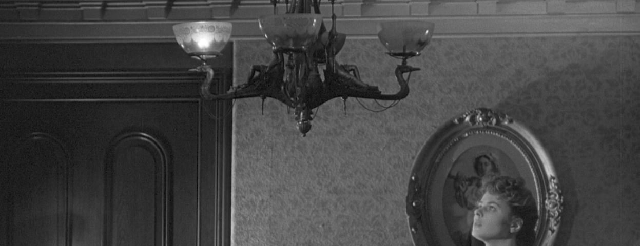 A black and white still from the 1944 film Gaslight. Paula, played by Ingrid Bergman, stares up with a haunted expression at a flickering gaslight chandelier in her home. Just one of its light is on.