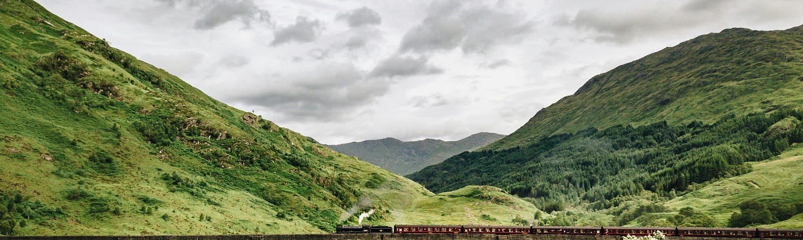 Steam train on a bridge with a Scottish mountainous, green landscape in the background.