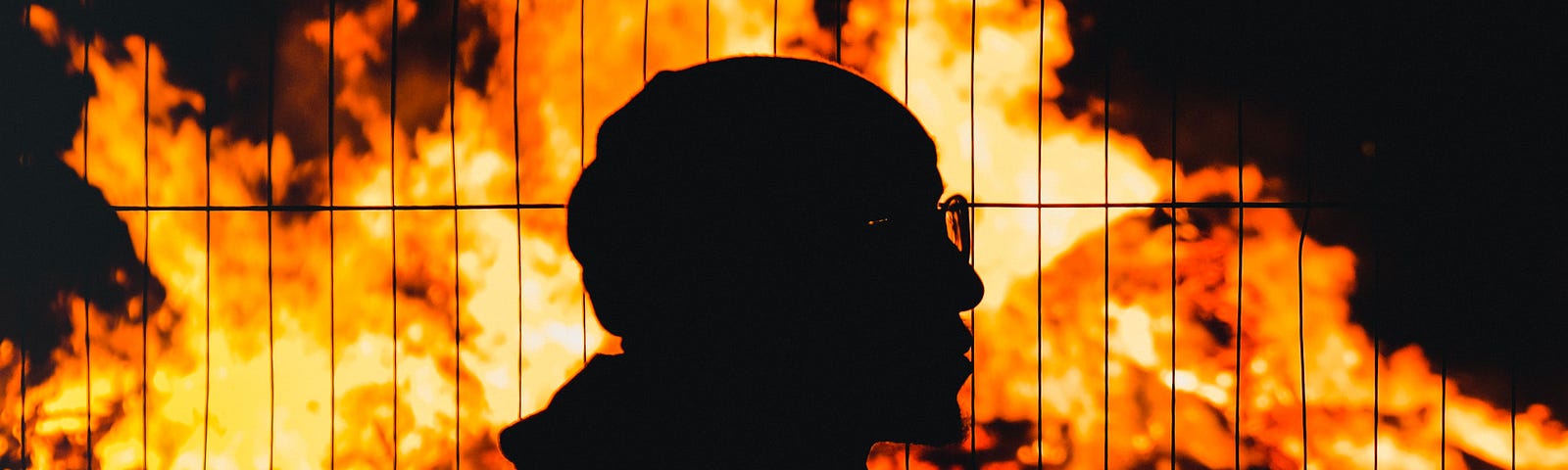 The silhouette of a bearded ethnic man in profile against a raging red burning fire behind a wired fence as if in protest.