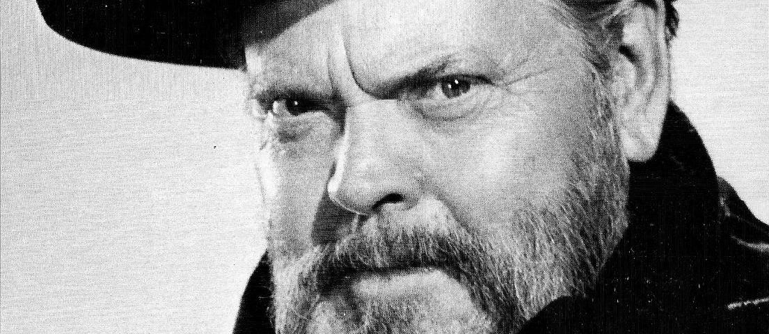 A black and white photograph of Orson Welles. He is wearing a thick black coat and scarf and a wide-brimmed black hat. He is older, late 50s, with a greying beard. He is looking directly at the camera with a smouldering expression. The photo is signed by the actor.