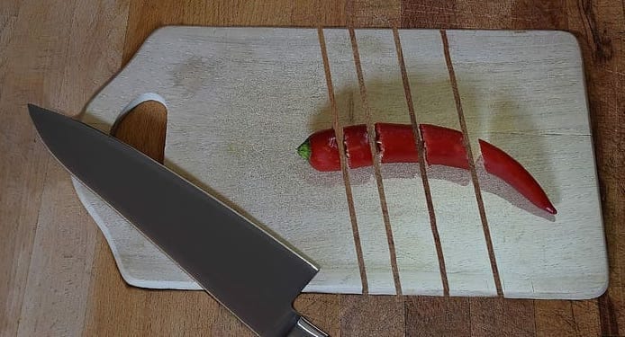 A knife resting on a cutting board with a chili pepper and lines cut through board.