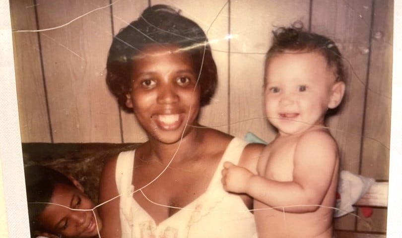 A Black woman smiling, looking straight into the camera while sitting on a couch with a pale skin baby in her left arm and a boy with a darker complexion sitting on her right, smiling and looking over at the baby.
