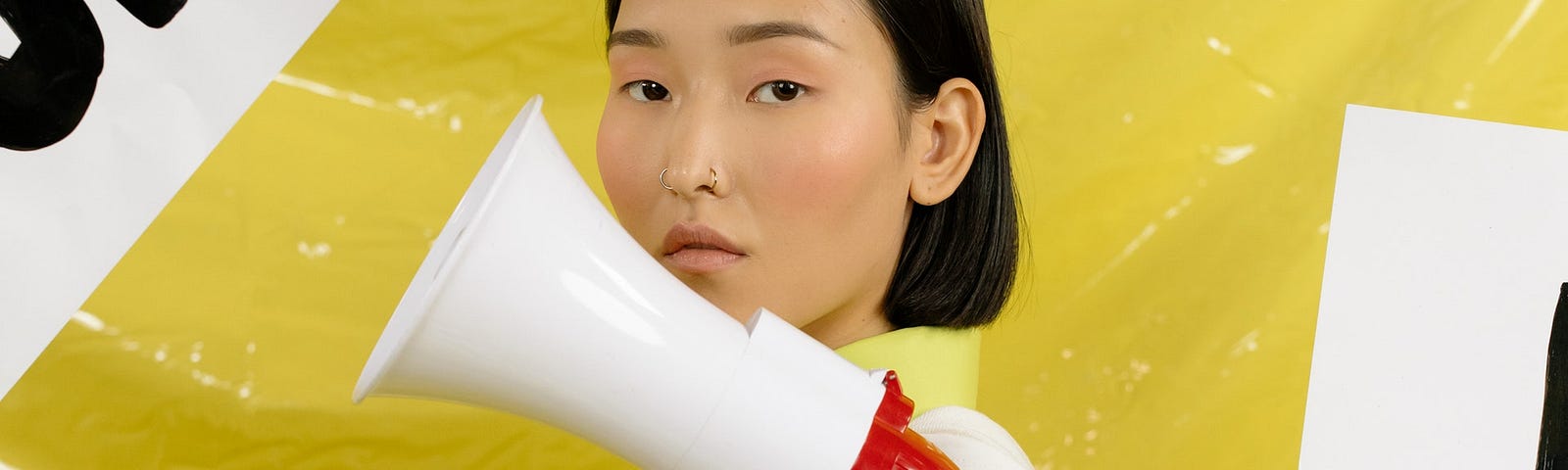 Woman with black, shoulder length hair, standing in front of a yellow backdrop, with posters saying, ‘don’t’ and ‘please stop’. The woman is holding a megaphone.