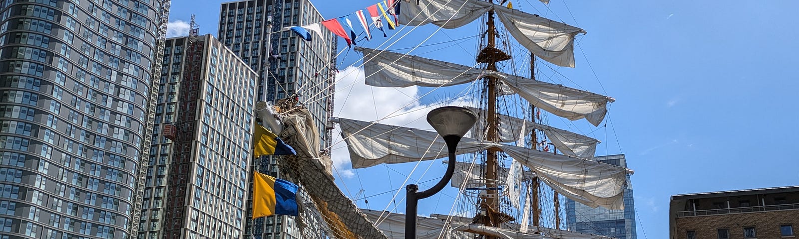 tall ship with tied-up sails in a dock with high-rise buildings covered in windows in background