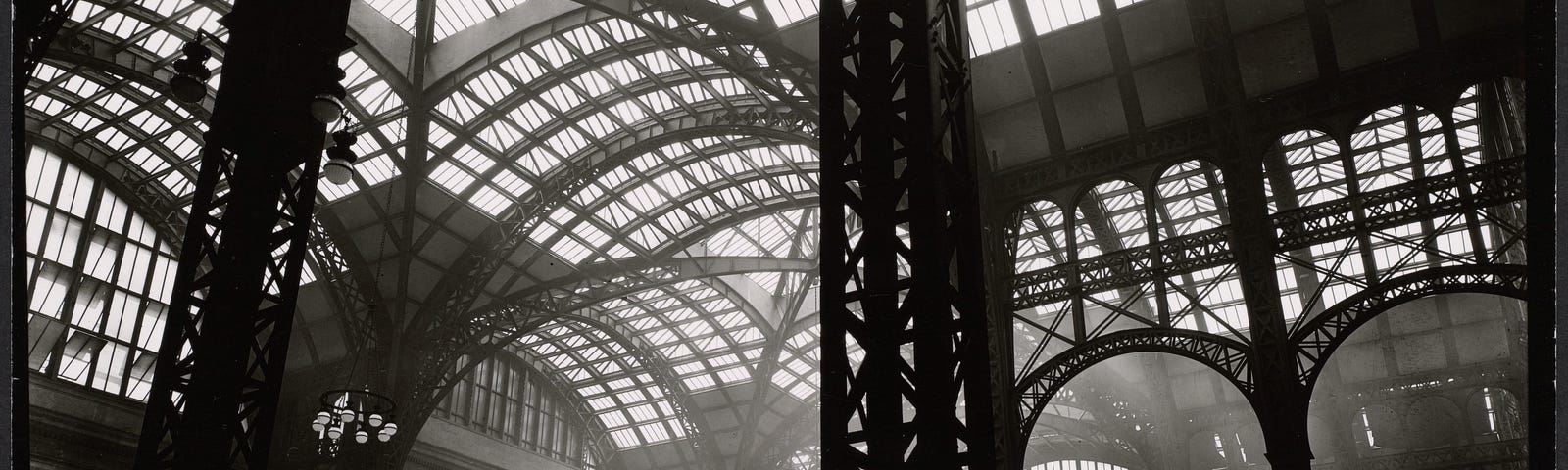 Massive steel uprights in the center and to the left, with lighter steel tracery and windows above; men stand near stairway to trains. Citation/Reference: CNY# 143–1 | Penn Station, Interior, Manhattan. Date 1935–1938, printed 1935-ca. 1990 |  Source https://digitalcollections.nypl.org/items/510d47d9-4e71-a3d9-e040-e00a18064a99 | Author Berenice Abbott (1898–1991) | Public Domain